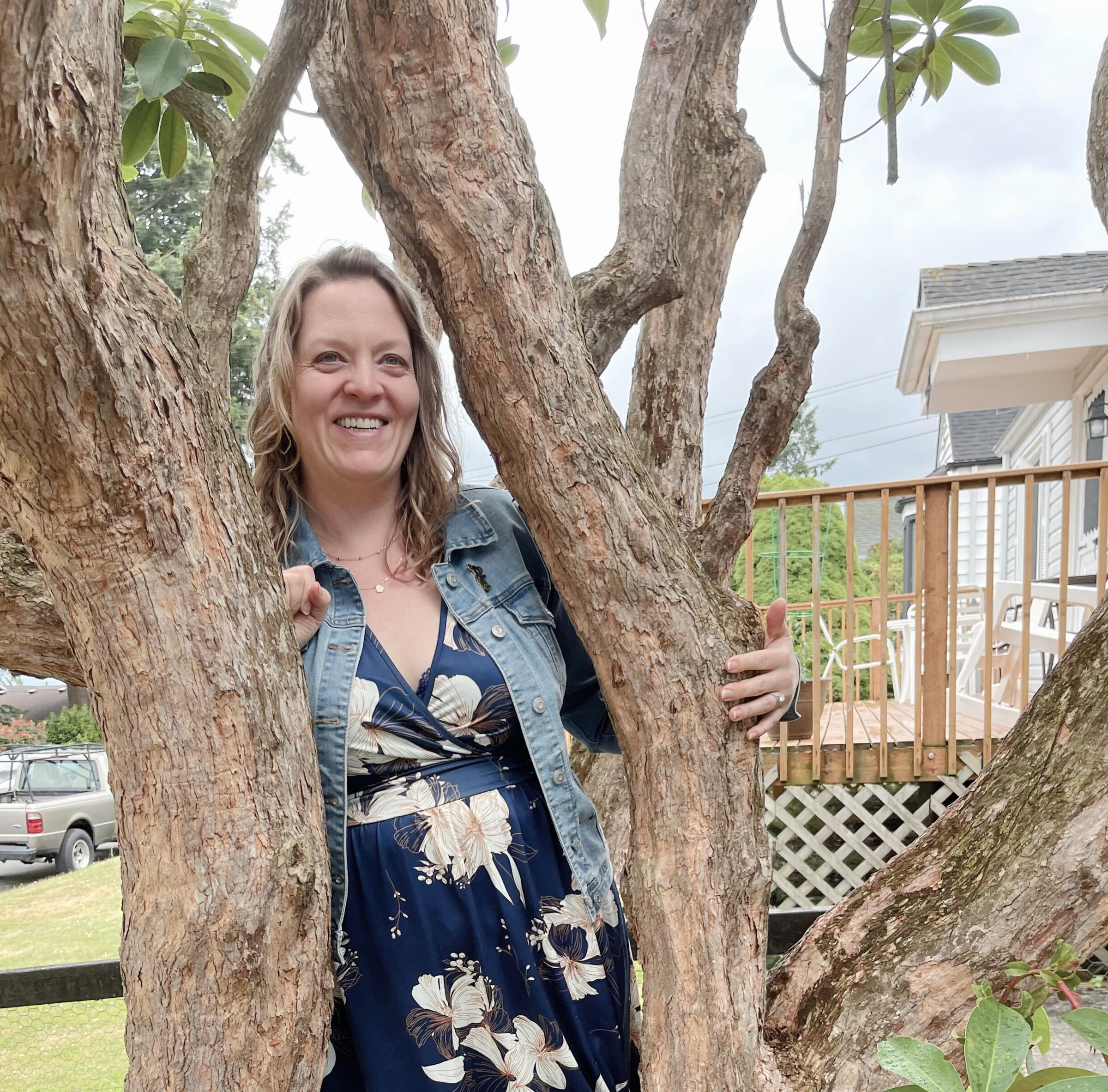 An image of a woman in a floral printed dress, she stands between the trunks of a tree, smiling.
